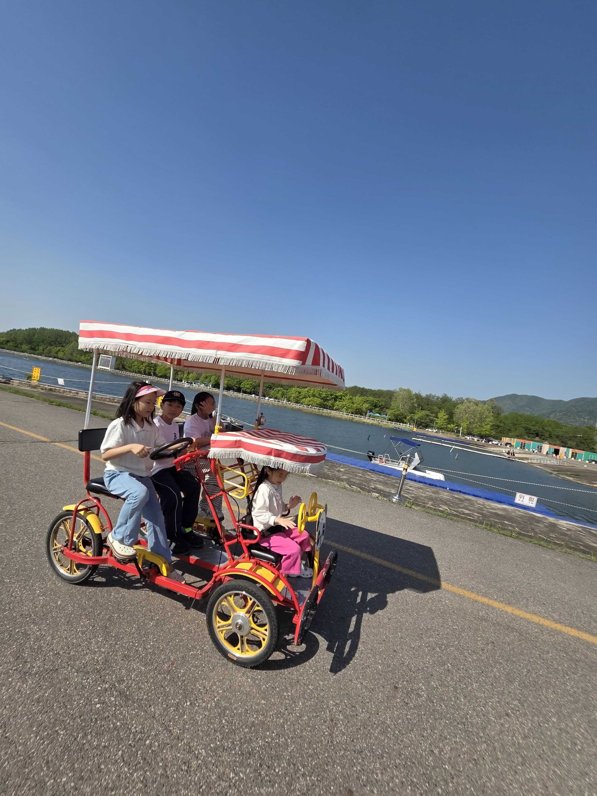 Misari Park Hanam four-seater pedal car with striped canopy on the riverside path with mountains in the background
