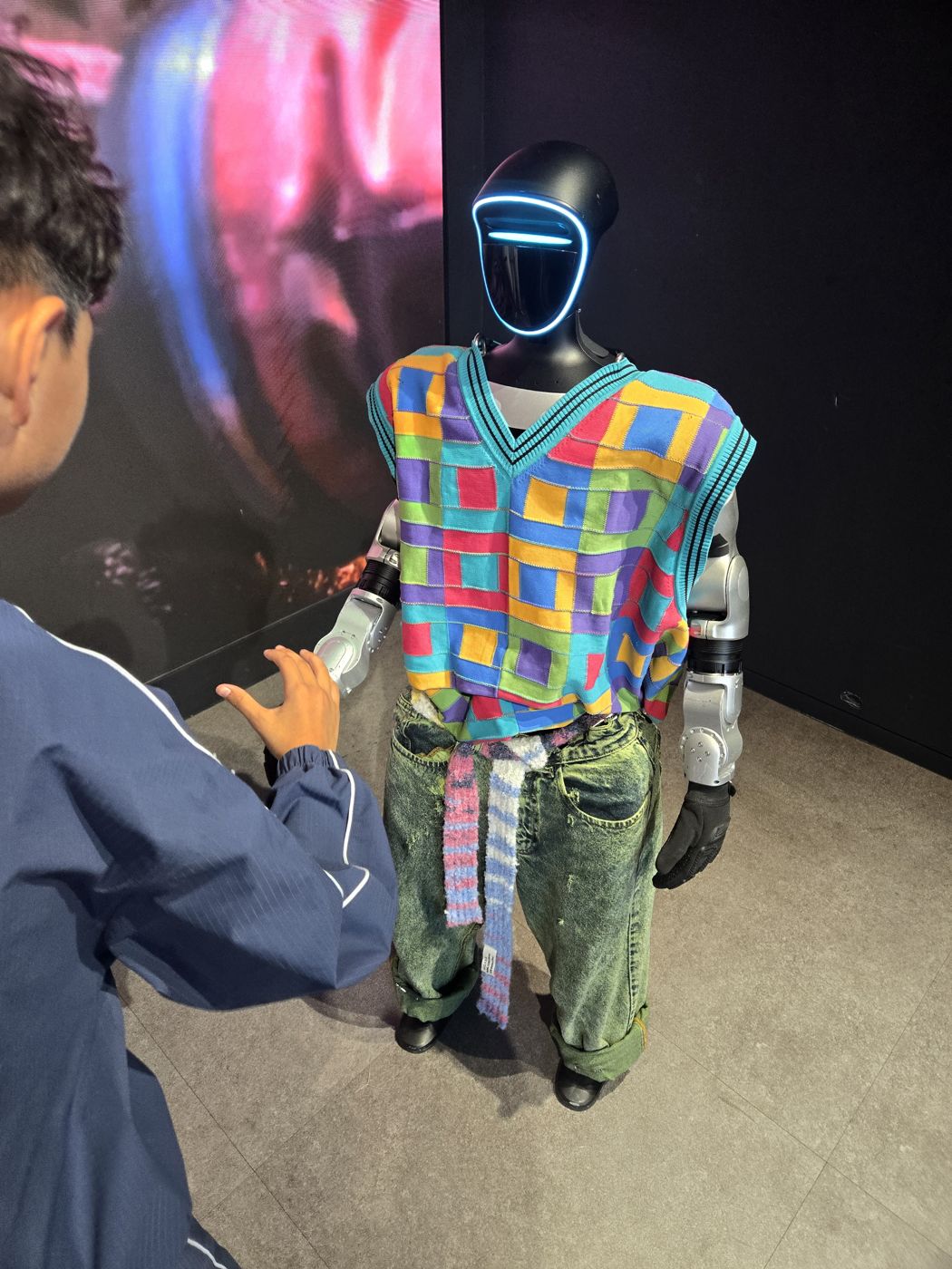Boy seen from behind reaching toward a humanoid robot in a colorful knit vest