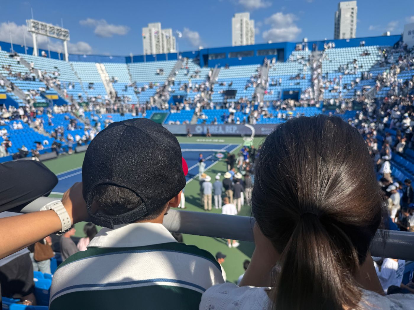 Two children seen from behind, a boy in a black cap and a girl with a ponytail, watching the court