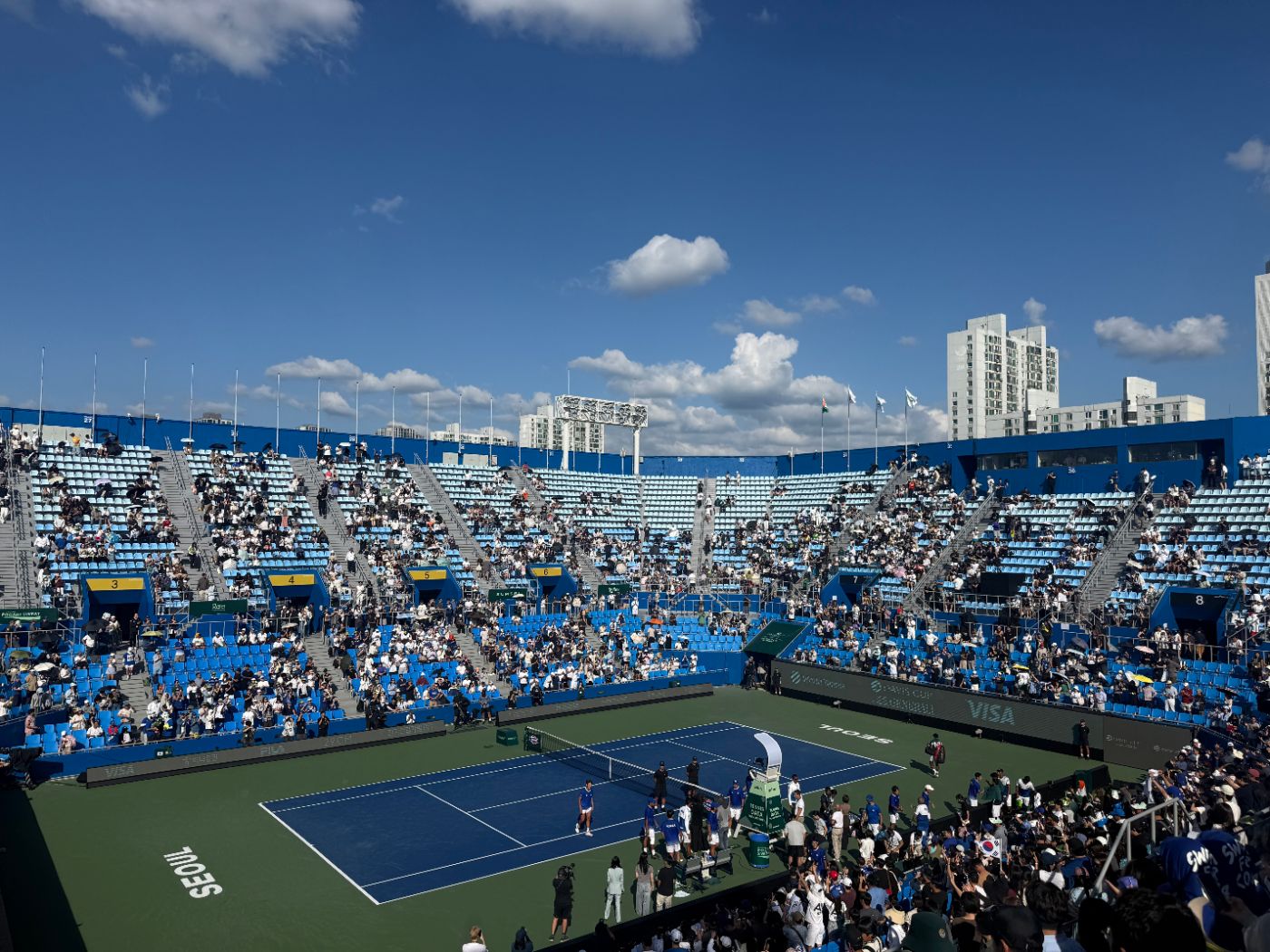 Wide view of Olympic Tennis Court under blue sky, with the Korean team walking onto the court
