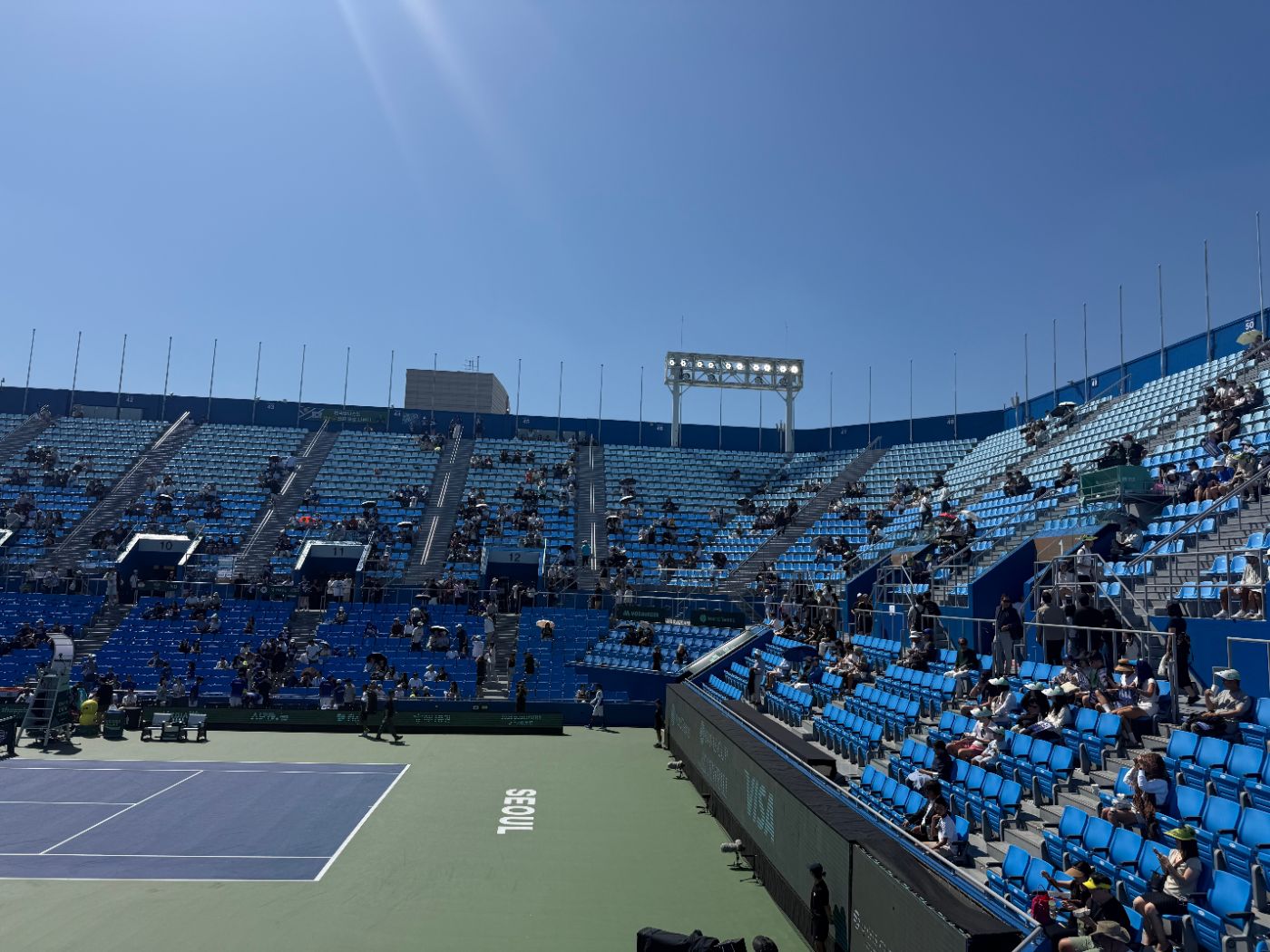 Lower seats beside the court in bright midday sun, with a few spectators under umbrellas